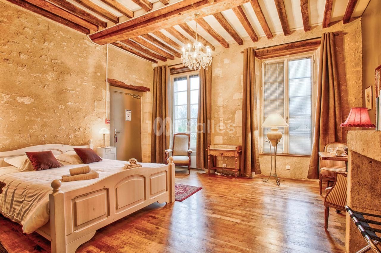 Chambre lumineuse avec lit double, poutres apparentes, murs en pierre, parquet et mobilier classique.