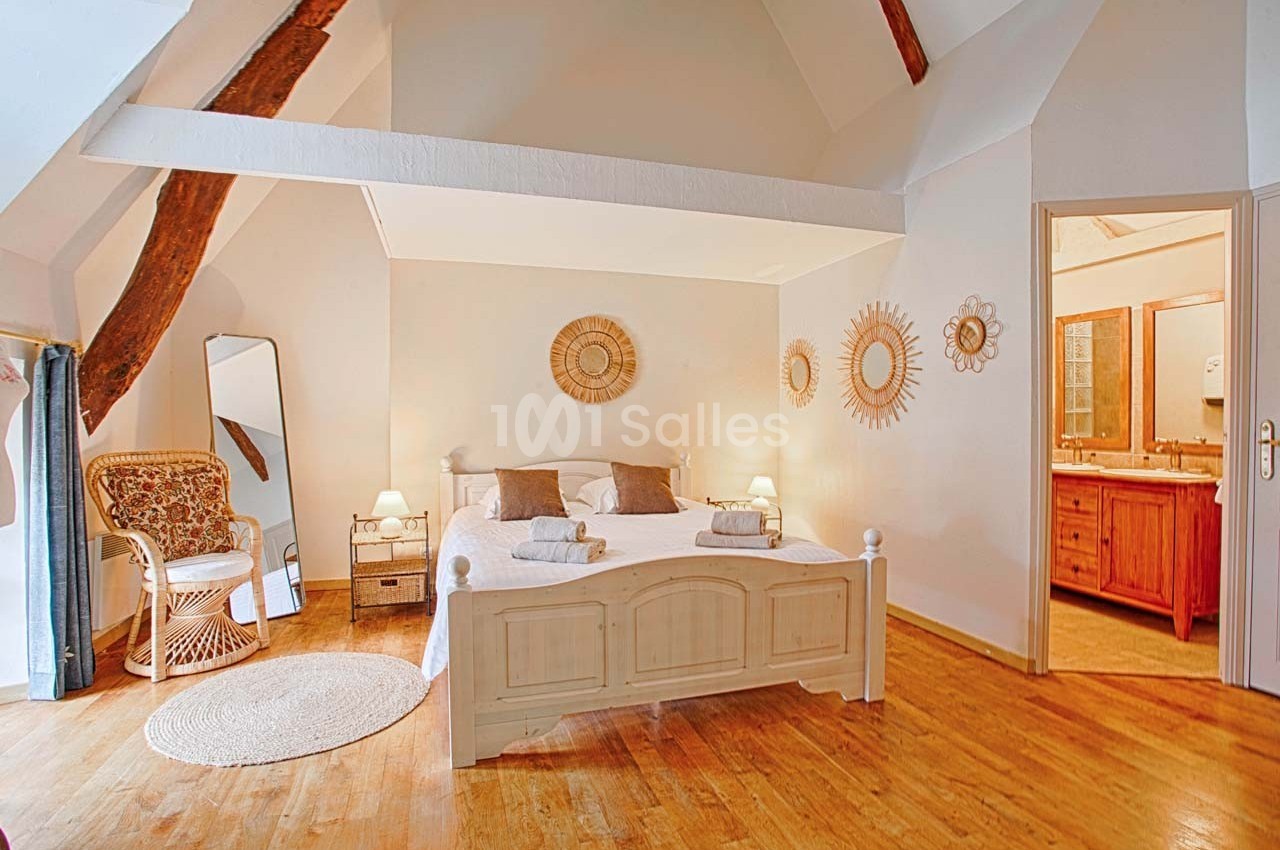 Chambre lumineuse avec lit double, poutres apparentes, miroir, fauteuil en osier et salle de bain attenante.