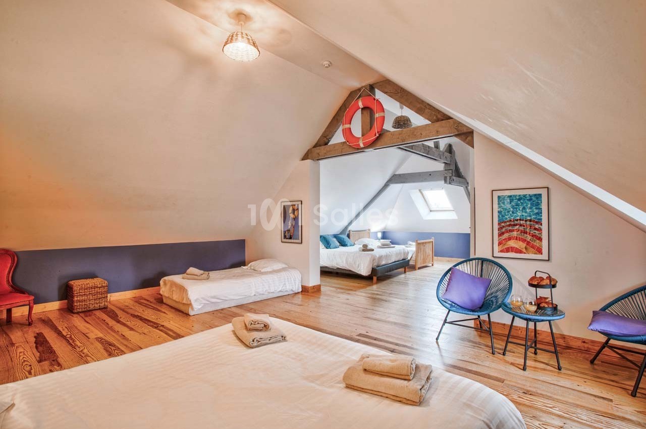 Chambre mansardée avec lits, fauteuils, table basse et décoration nautique, éclairée par une fenêtre de toit.