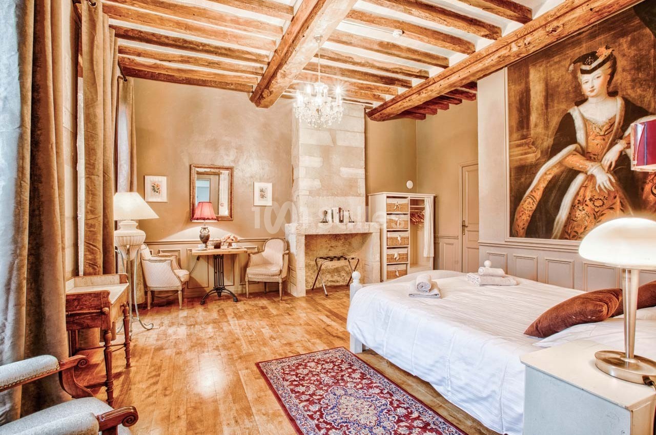 Chambre lumineuse avec poutres apparentes, lit double, coin bureau, cheminée en pierre et grand tableau mural.