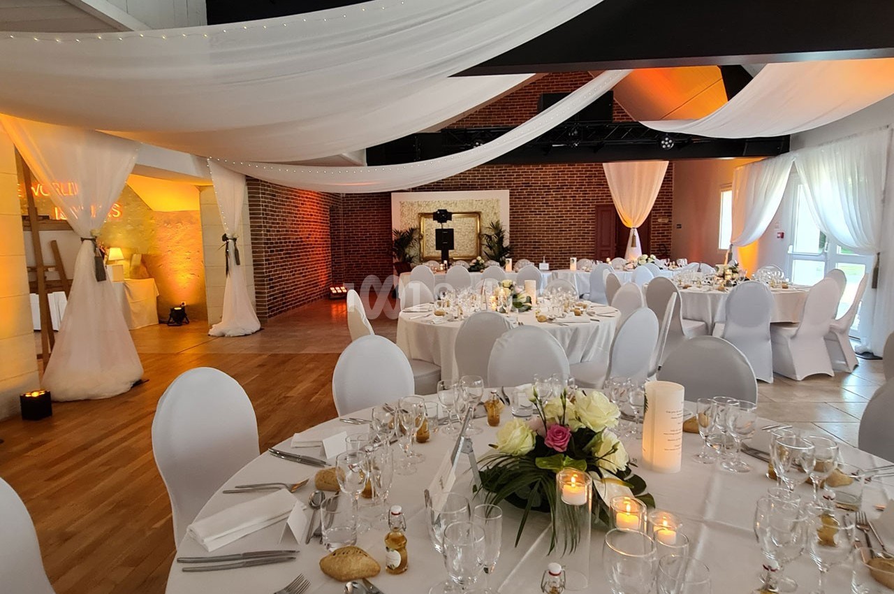 Salle de réception décorée avec des tables rondes dressées, des chaises blanches et des voilages suspendus.