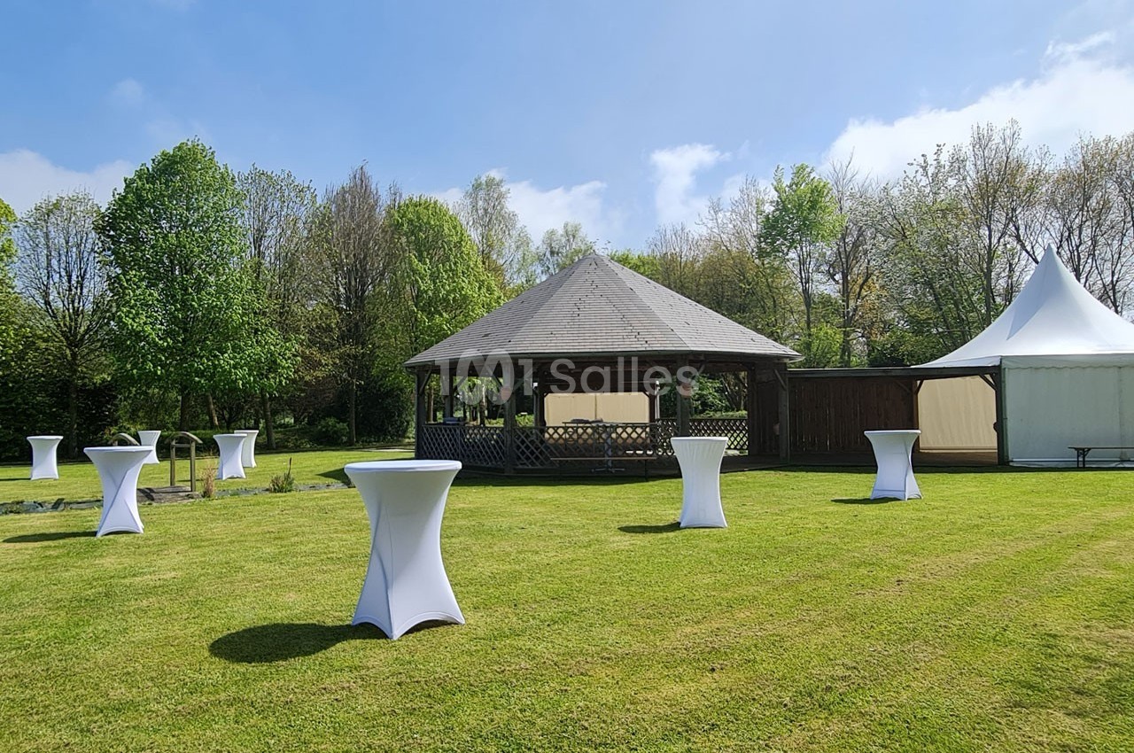 Espace extérieur avec pelouse, tables hautes couvertes de nappes blanches, kiosque en bois et tente blanche.