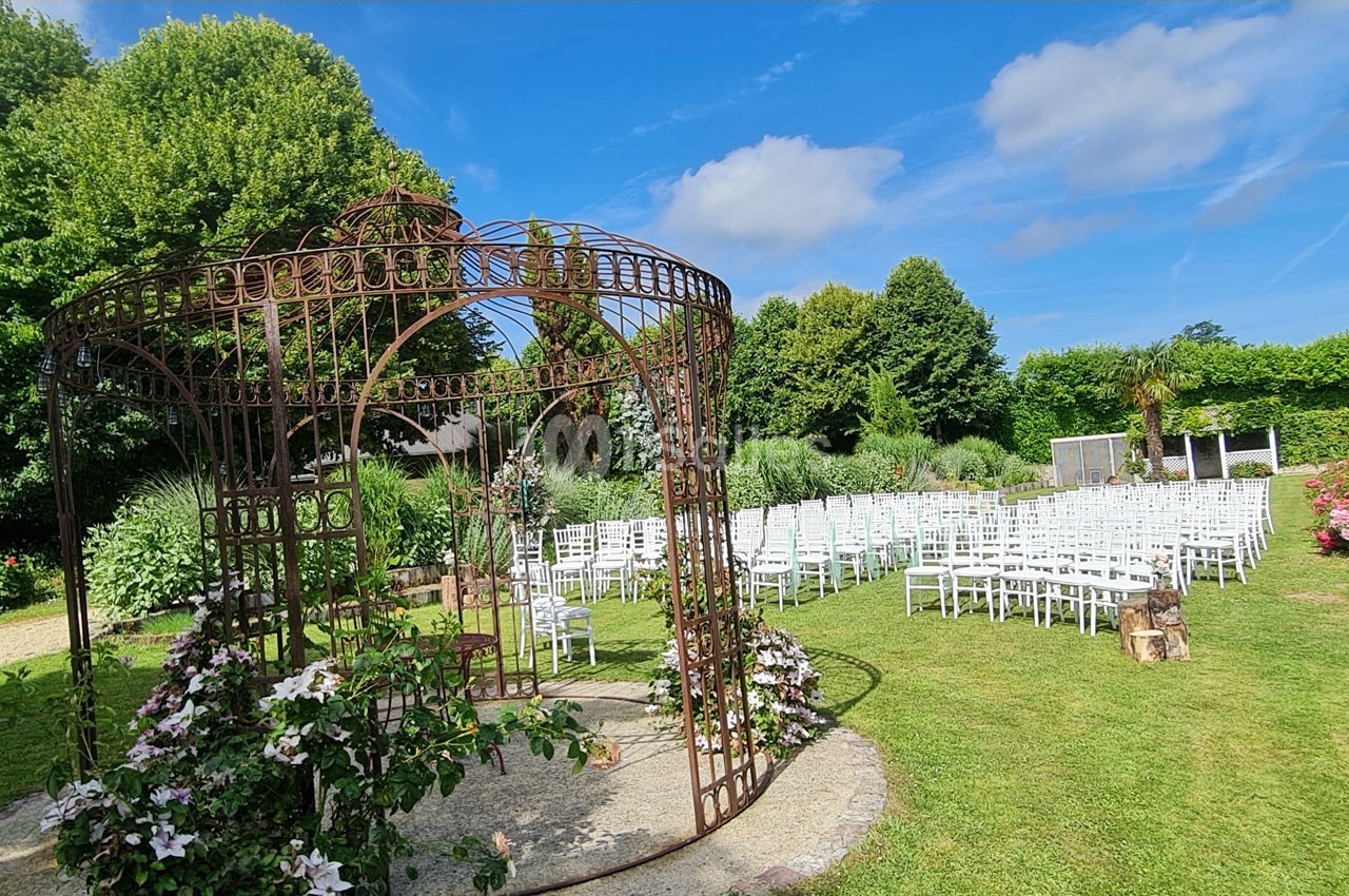 Structure métallique décorative entourée de fleurs dans un jardin, avec des rangées de chaises blanches en arrière-plan.