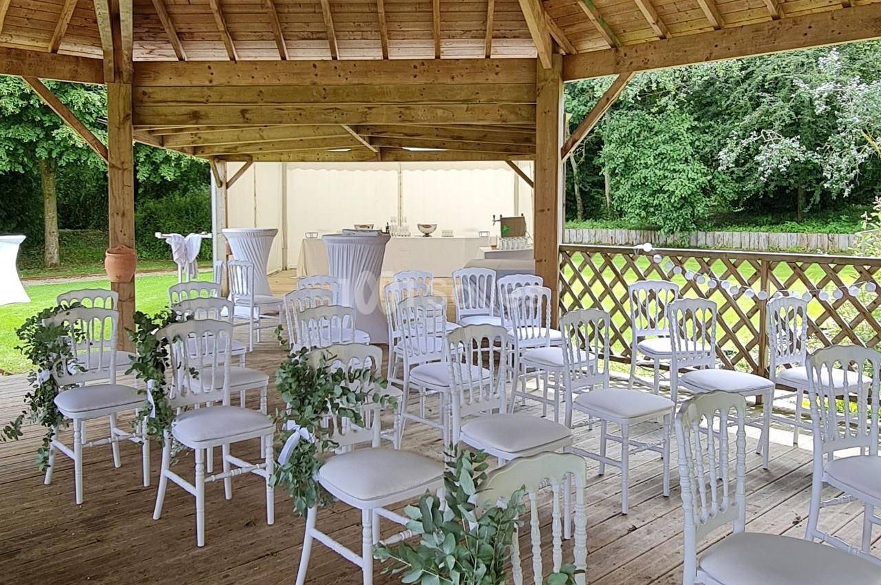 Chaises blanches décorées de feuillage disposées sous une pergola en bois, entourée de verdure.