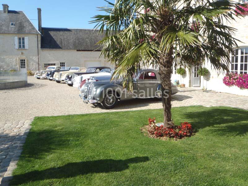 Plusieurs voitures anciennes stationnées dans la cour d'une bâtisse en pierre, avec pelouse et palmier au premier plan.