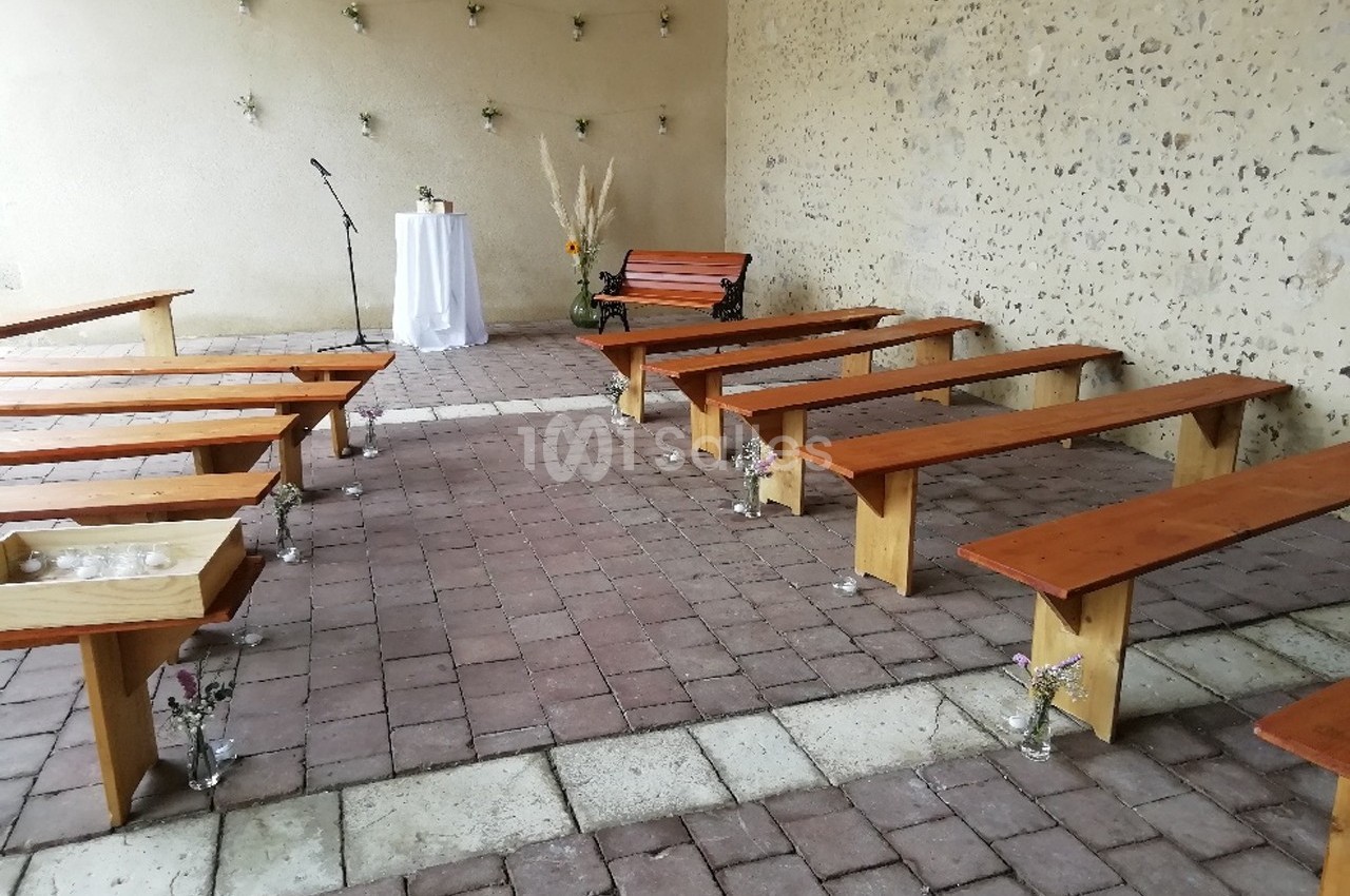 Bancs en bois disposés dans une cour pavée avec un pupitre, une table décorée et des fleurs en arrière-plan.