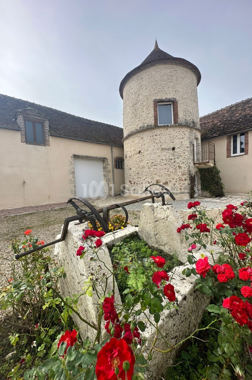 Vieille bâtisse en pierre avec une tour ronde, entourée de fleurs rouges dans une cour pavée.