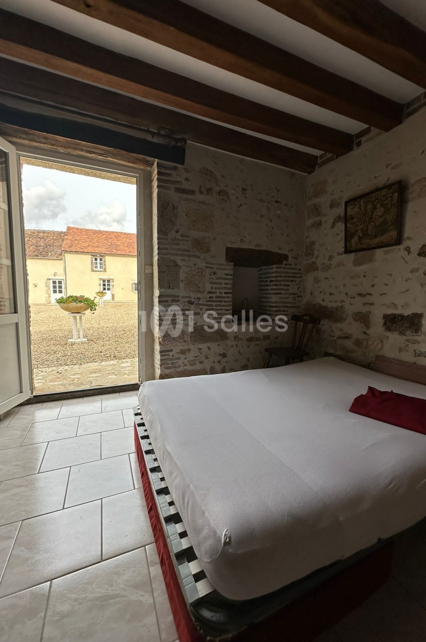 Chambre avec lit double, murs en pierre, poutres apparentes, et porte ouverte sur une cour pavée avec bâtiments anciens.