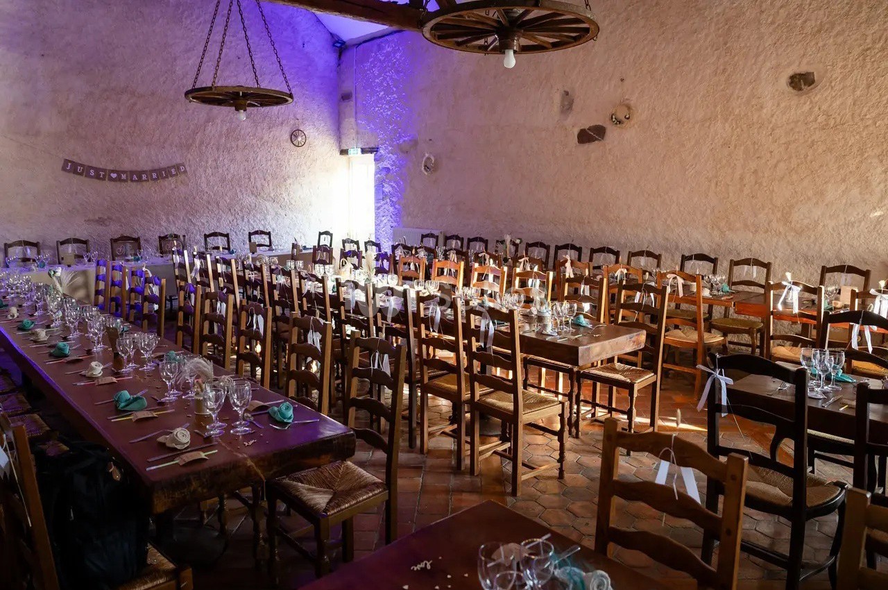 Salle de réception rustique avec de longues tables en bois dressées, entourées de chaises, éclairée par une lumière douce.