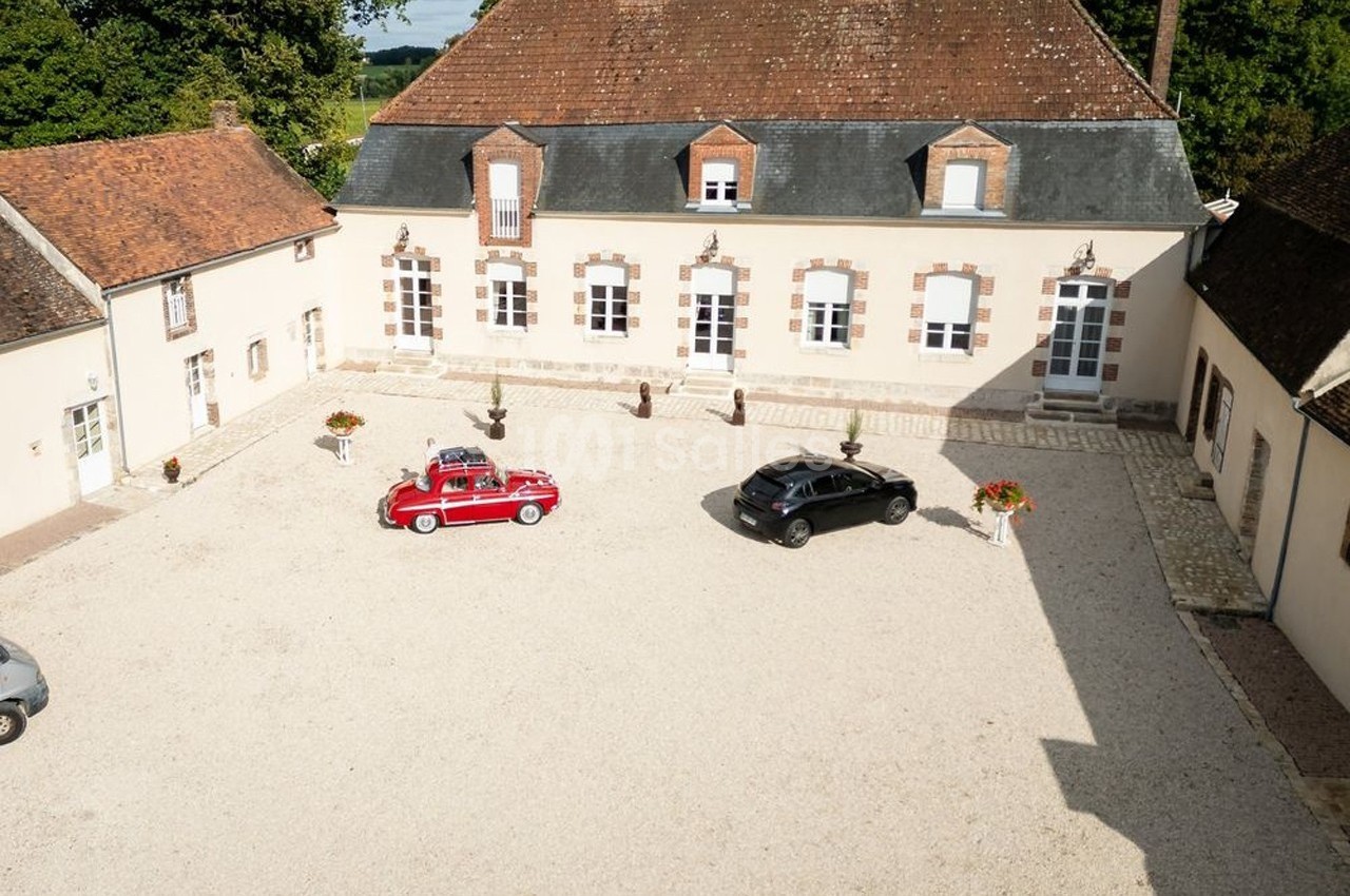 Cour d'une grande maison en pierre avec toit en tuiles, deux voitures garées, une rouge et une noire.