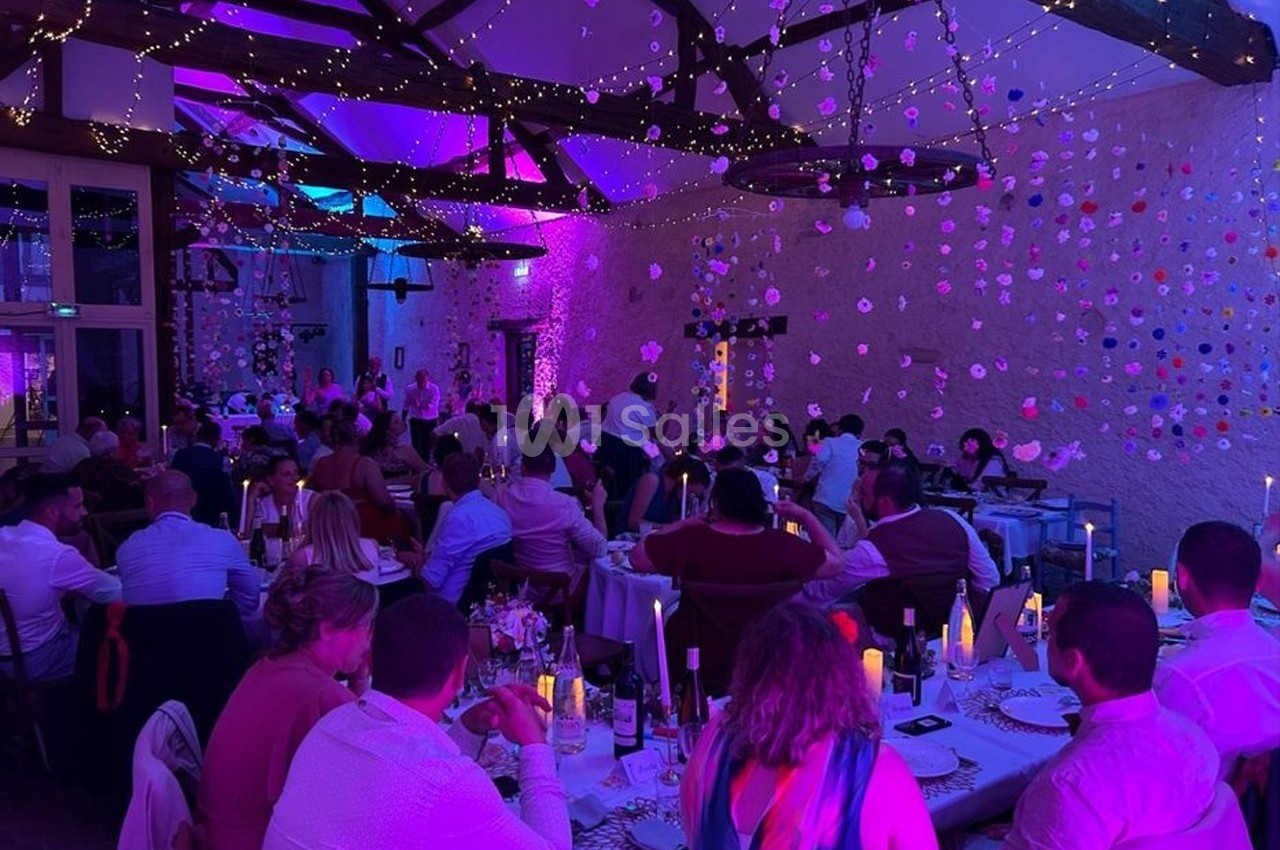 Salle de réception décorée avec guirlandes lumineuses et fleurs suspendues, des invités dînent à des tables rondes.