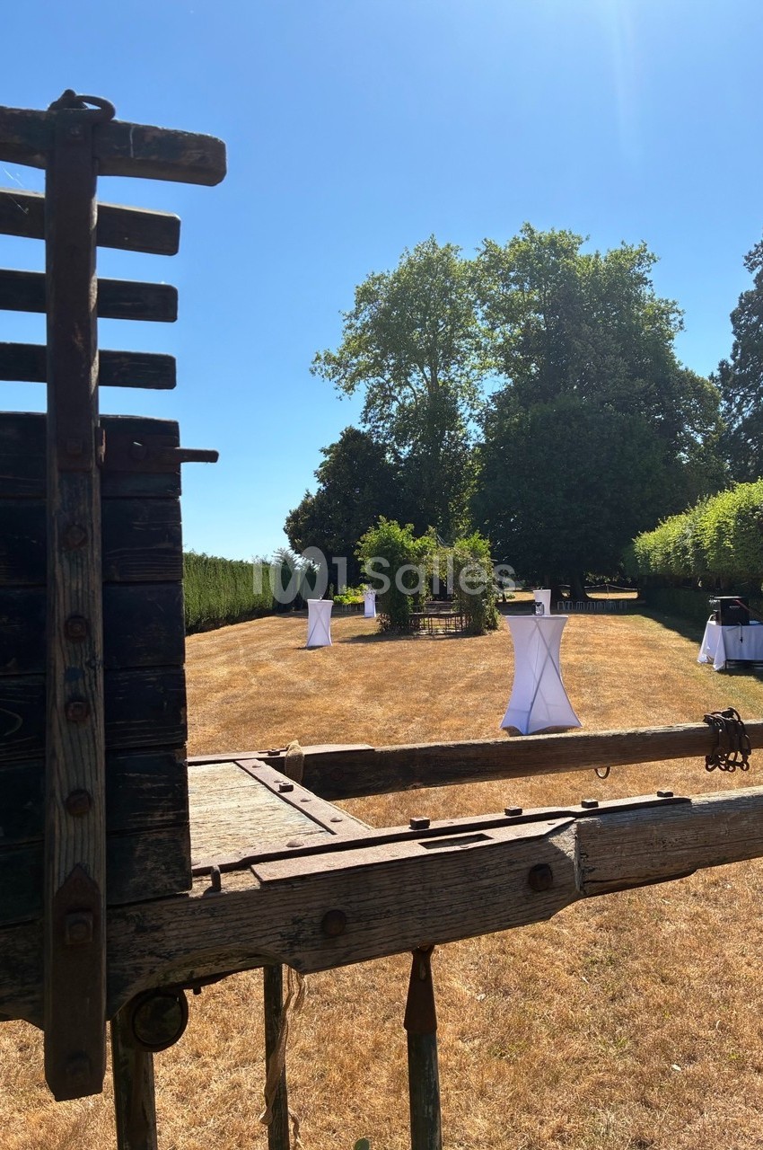 Vue d'un jardin ensoleillé avec des tables hautes drapées de blanc, entouré de verdure et d'arbres.