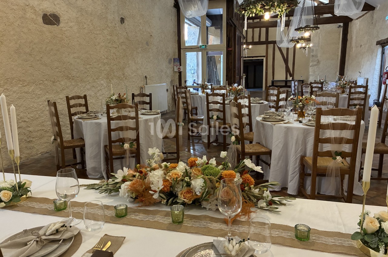 Salle dressée pour mariage avec tables rondes, chaises en bois, nappes crème et décoration florale abricot-blanc.