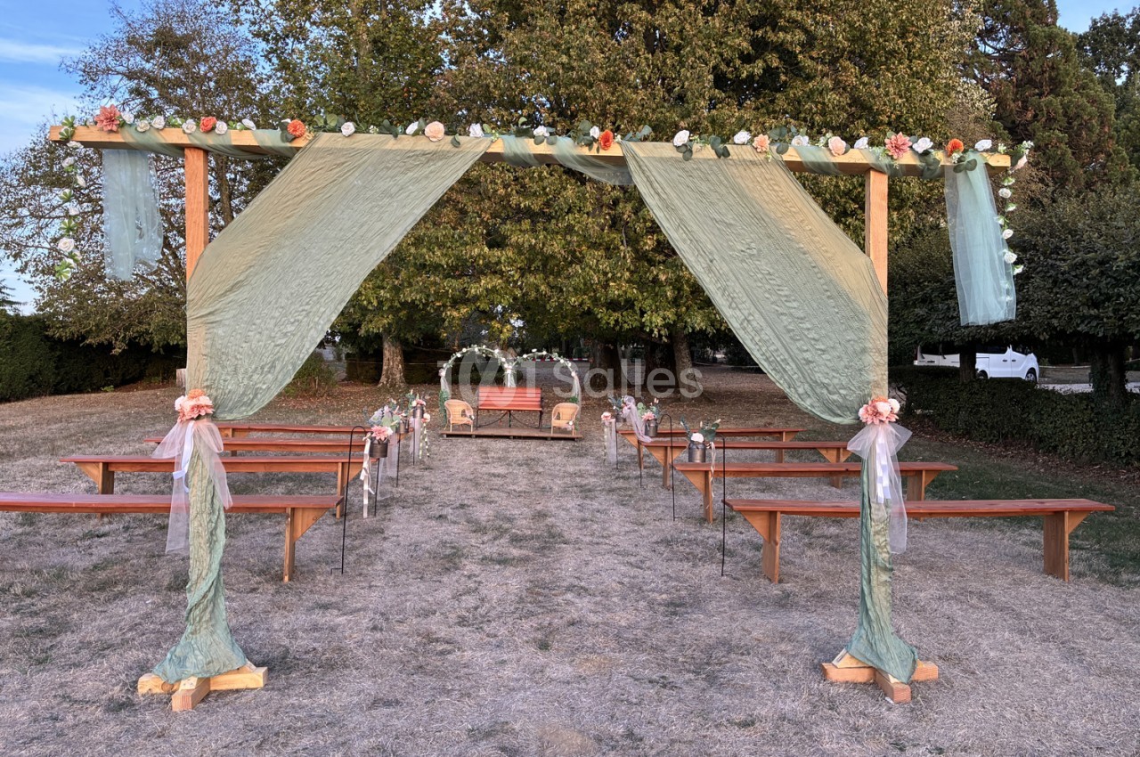 Cérémonie en plein air sous pergola en bois décorée de voiles et fleurs roses, bancs en bois de chaque côté.