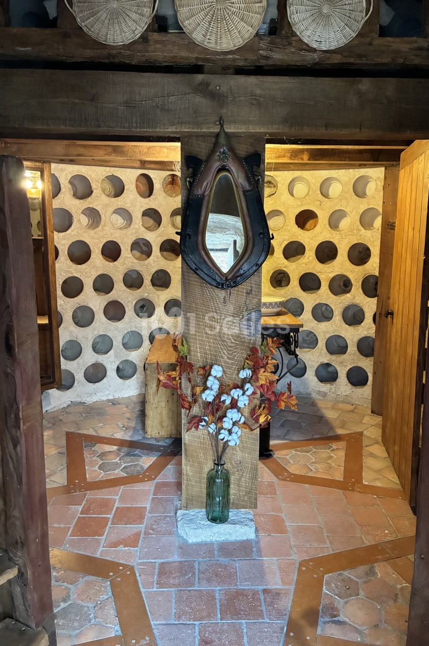 Intérieur rustique avec sol en tomettes, murs en pierre percés de trous circulaires, miroir en fer forgé et vase de fleurs.