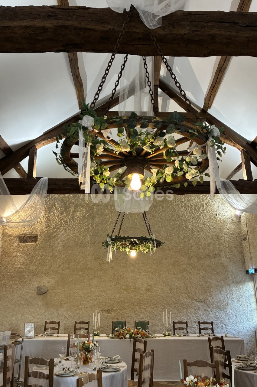 Salle voûtée en pierre avec poutres apparentes, tables dressées en blanc et chaises rustiques, décorée de guirlandes florales suspendues.