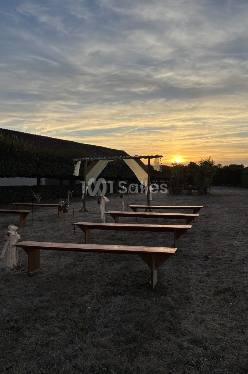 Espace extérieur avec tables de bois alignées, tonnelle blanche et coucher de soleil sur colline boisée.