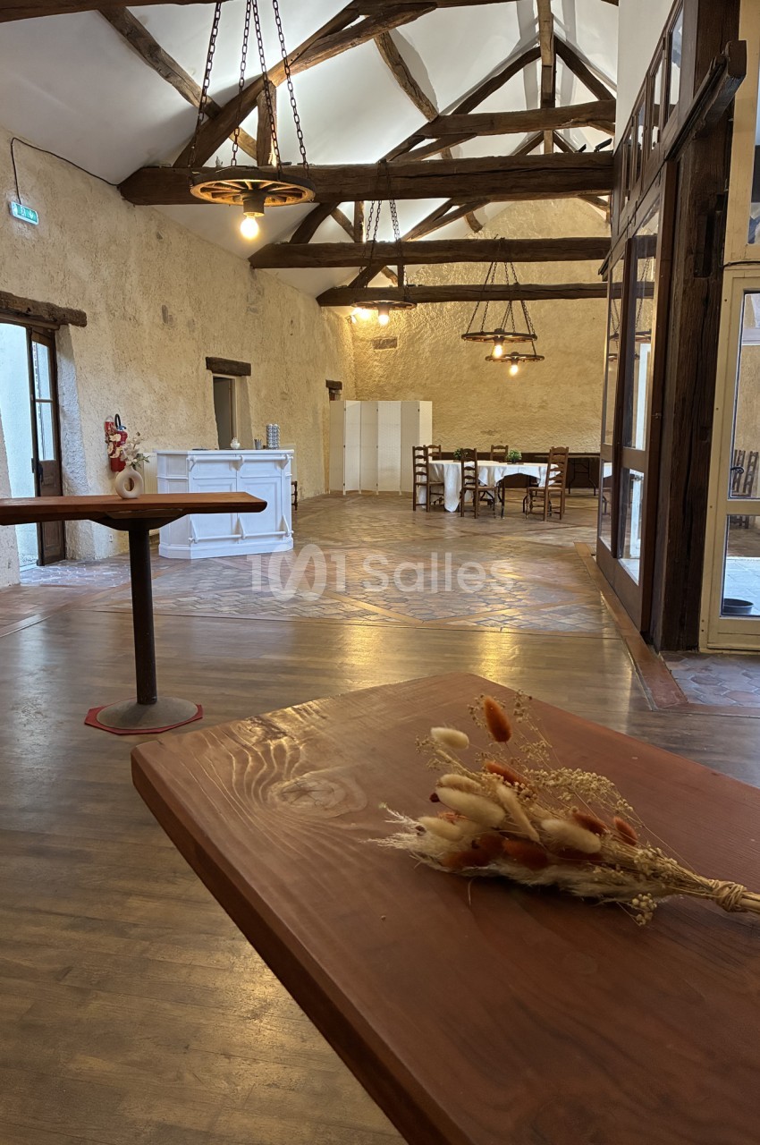 Salle avec poutres apparentes, murs pierre, sol parquet et luminaires suspendus, table dressée au fond.