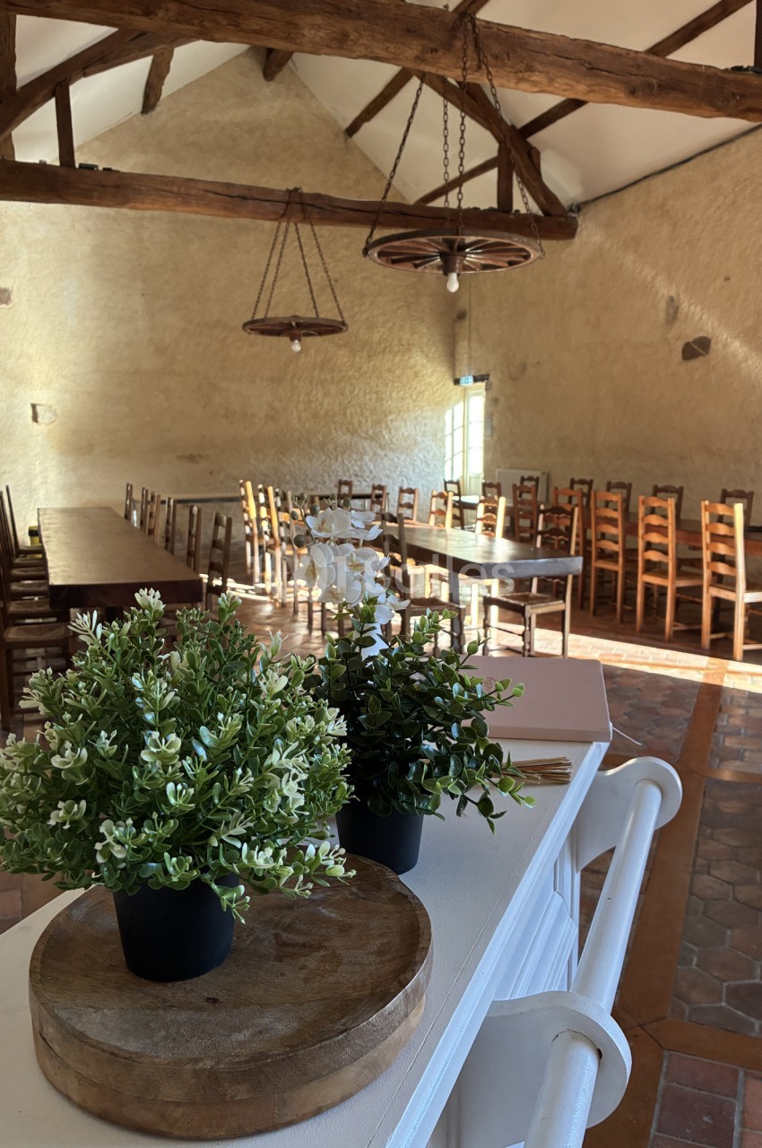 Salle intérieur dressée avec tables longues, chaises en bois, poutres apparentes et lustre style rustique.