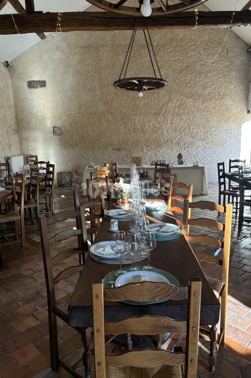 Salle voûtée en pierre avec poutres apparentes, tables dressées et chaises en bois, luminaire suspendu.