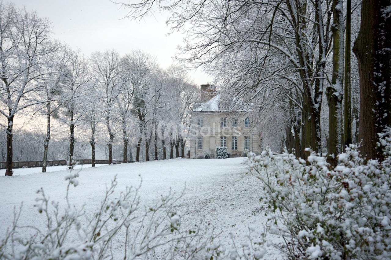 Manoir entouré d'arbres et d'un jardin recouverts de neige en hiver, sous un ciel nuageux.