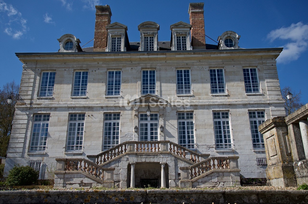 Façade d'un manoir en pierre avec escaliers symétriques, grandes fenêtres et cheminées, sous un ciel dégagé.