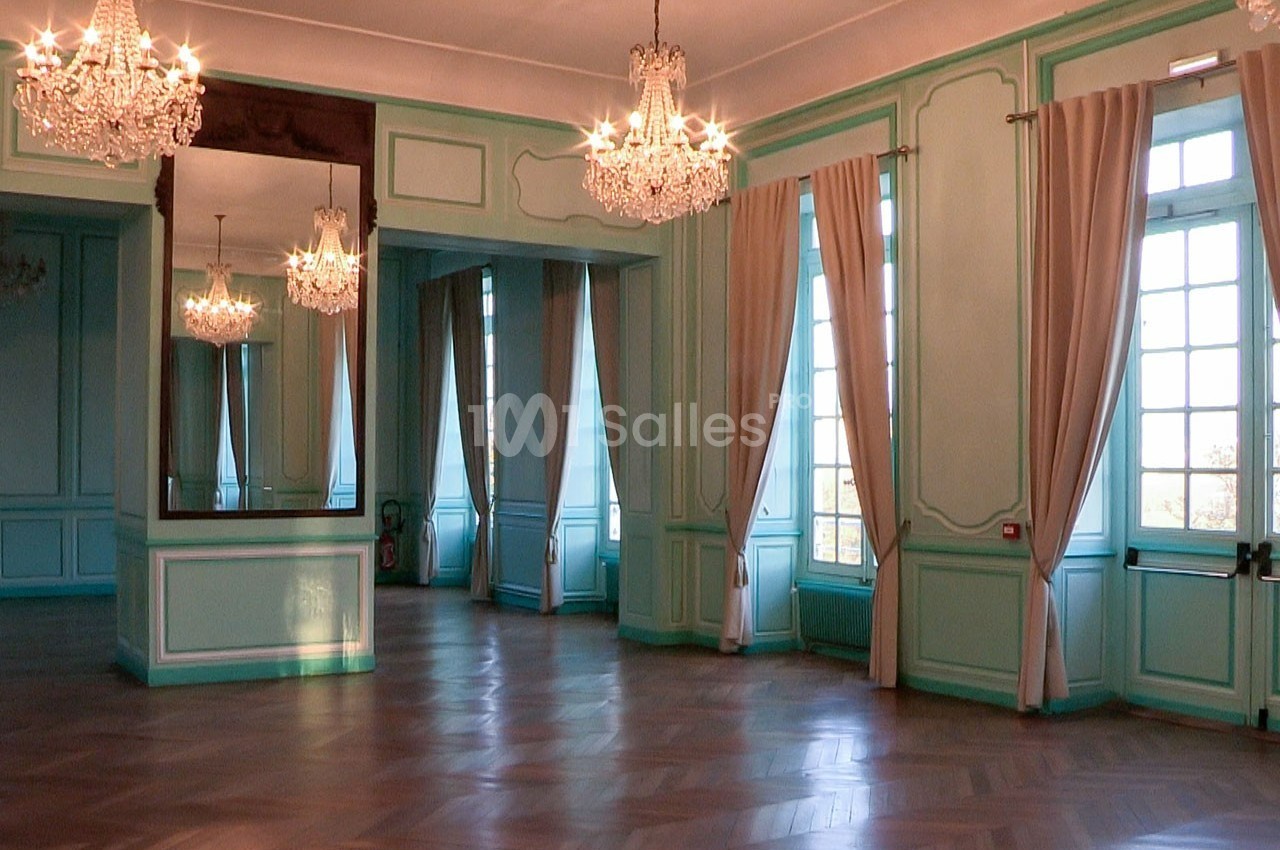 Salle lumineuse avec parquet en bois, murs turquoise, grands miroirs, lustres et rideaux beiges encadrant des fenêtres.