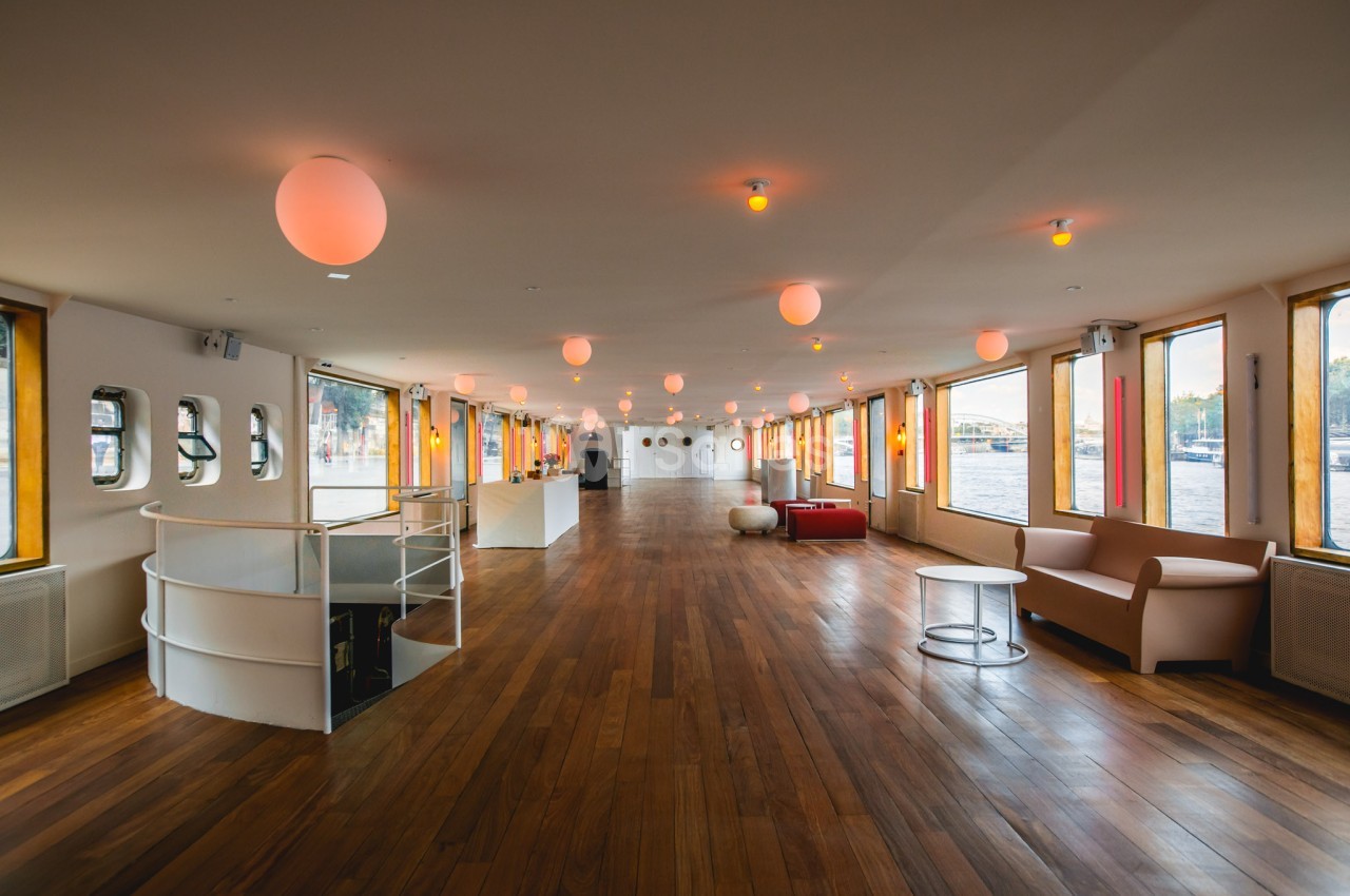 Salle spacieuse avec parquet en bois, canapés, baies vitrées et éclairage suspendu, offrant une vue sur l'extérieur.