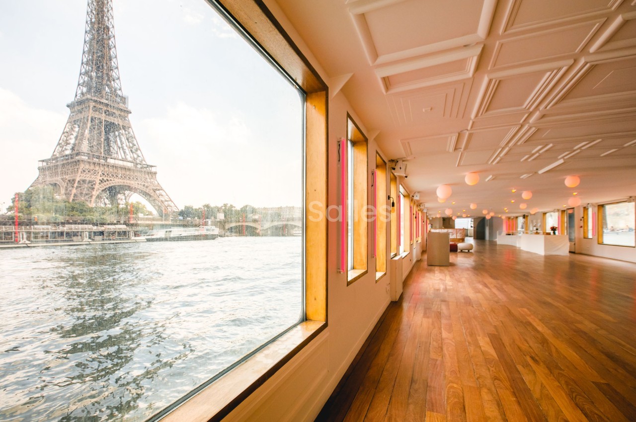 Vue intérieure d'une salle avec parquet et grandes fenêtres donnant sur la Tour Eiffel et la Seine.