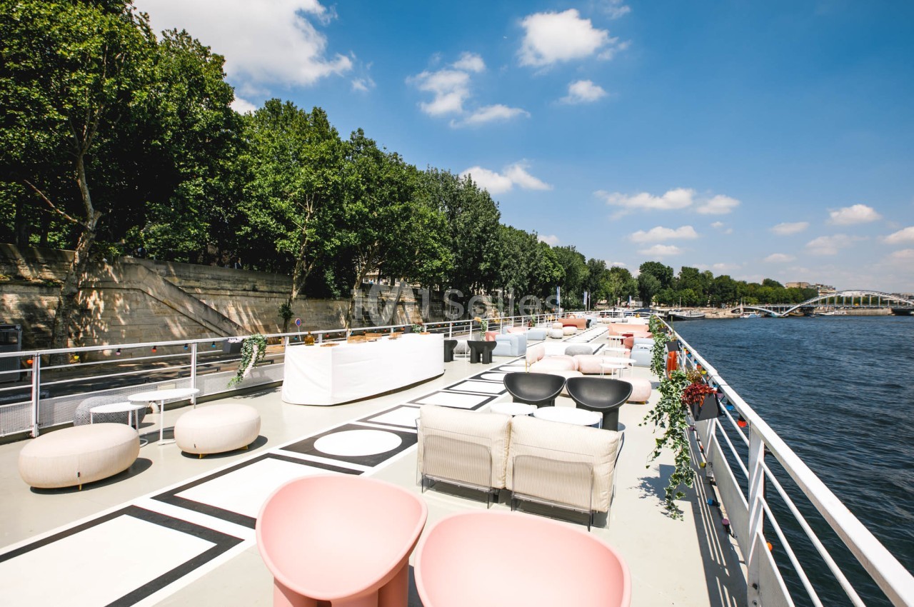 Terrasse aménagée sur une péniche avec mobilier moderne, bordée d'arbres et vue sur la Seine par temps ensoleillé.