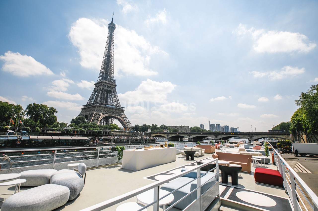 Terrasse aménagée sur une péniche avec vue sur la Tour Eiffel et la Seine par une journée ensoleillée.