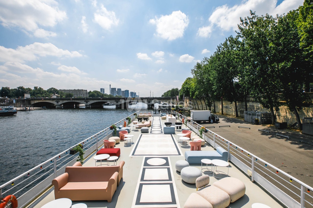 Terrasse aménagée sur un bateau avec vue sur une rivière, un pont, des arbres et des immeubles en arrière-plan.