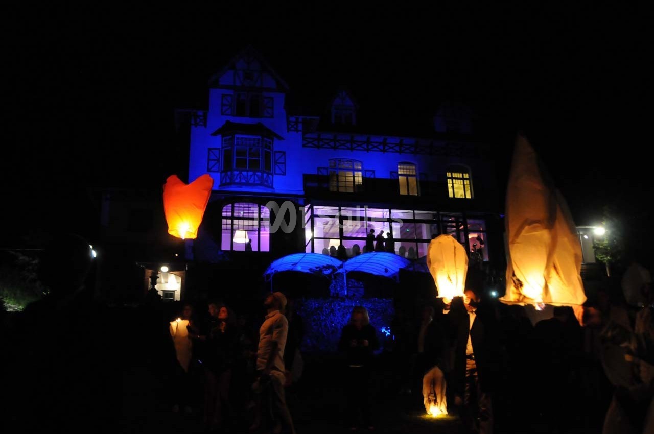 Des lanternes lumineuses flottent dans la nuit près d'une grande maison éclairée en bleu, entourée de personnes.