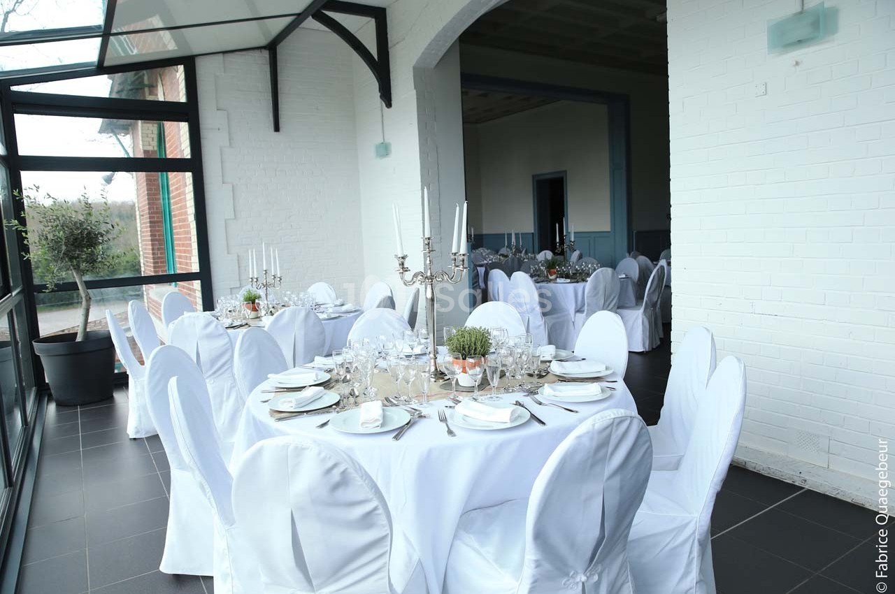 Salle lumineuse avec tables rondes dressées, nappes blanches, chandeliers et baies vitrées donnant sur l'extérieur.