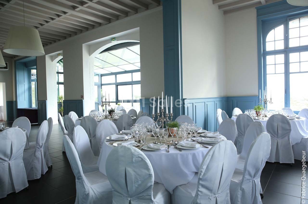 Salle de réception lumineuse avec tables rondes dressées, nappes blanches et chandeliers élégants.