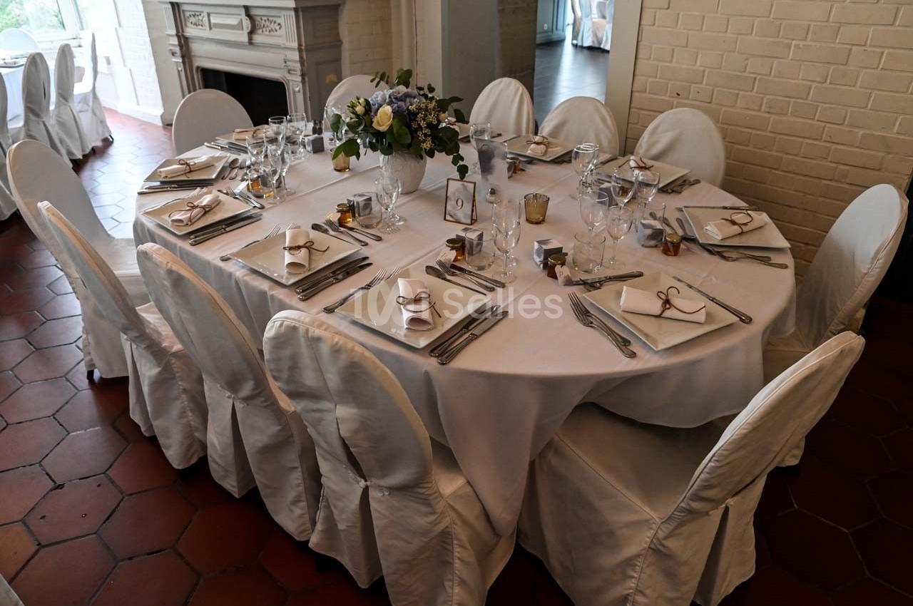 Table ronde dressée avec nappes blanches, vaisselle élégante, bouquets de fleurs et chaises couvertes.