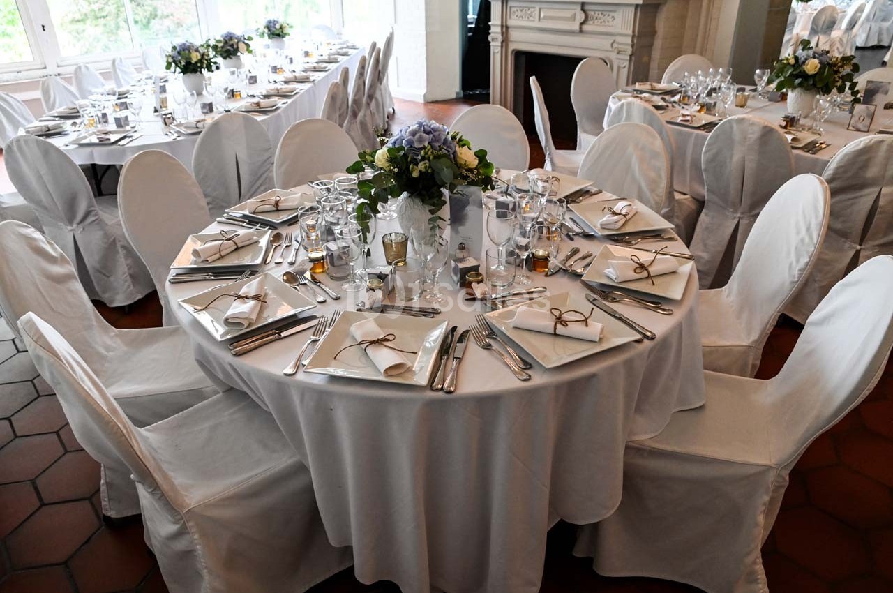 Table ronde élégamment dressée avec nappes blanches, couverts, verres et centre de table floral dans une salle lumineuse.