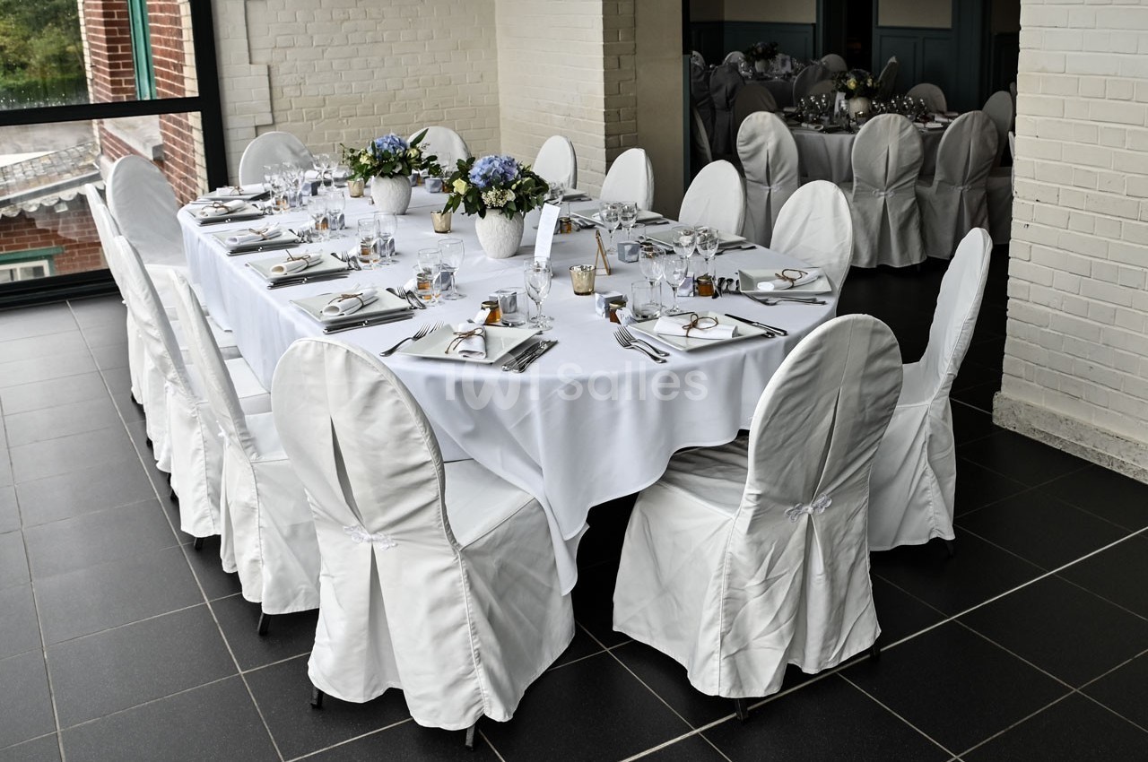 Table dressée pour un repas formel avec nappes blanches, chaises couvertes, vaisselle élégante et décorations florales.