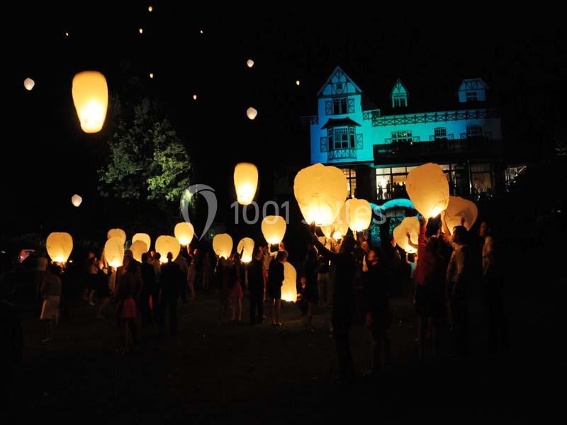 Des personnes lâchent des lanternes lumineuses dans le ciel nocturne devant un grand bâtiment éclairé.