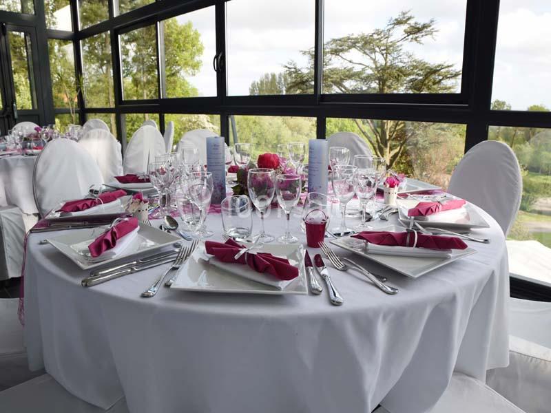 Table ronde élégamment dressée avec nappes blanches, serviettes roses pliées et vaisselle raffinée, près de grandes baies…