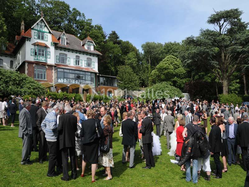 Groupe de personnes rassemblées dans un jardin devant une grande maison de style traditionnel, par une journée ensoleillée.