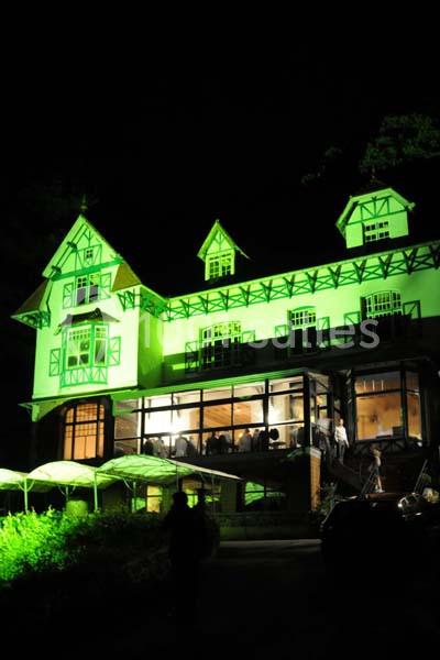 Maison à colombages éclairée en vert la nuit, avec des fenêtres illuminées et une terrasse partiellement visible.