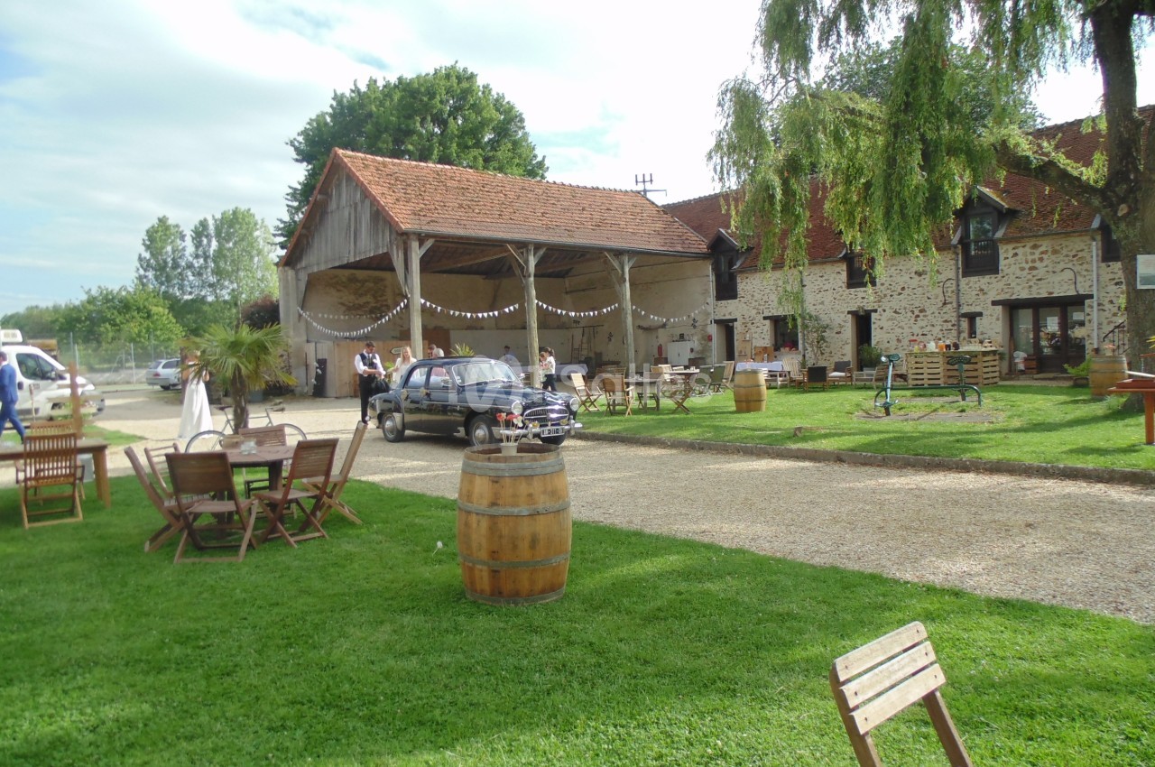 Cour extérieure d'une ferme rénovée avec tables, chaises, tonneaux décoratifs et une voiture garée sous un auvent.