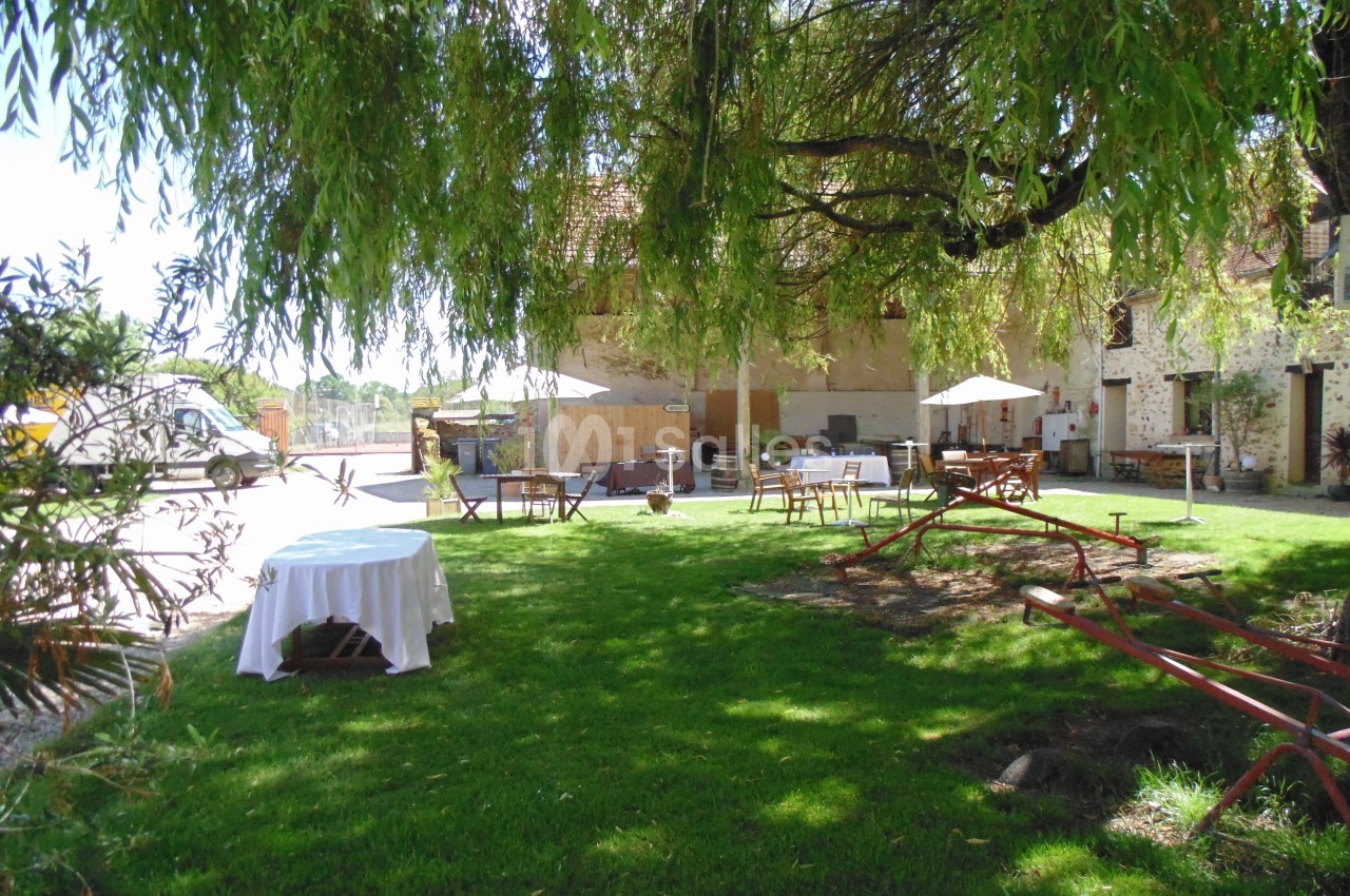 Jardin verdoyant avec tables, chaises et parasols sous un grand arbre, près d'un bâtiment en pierre.