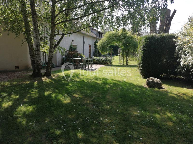 Jardin verdoyant avec pelouse, arbres, table et chaises près d'une maison blanche sous un ciel ensoleillé.