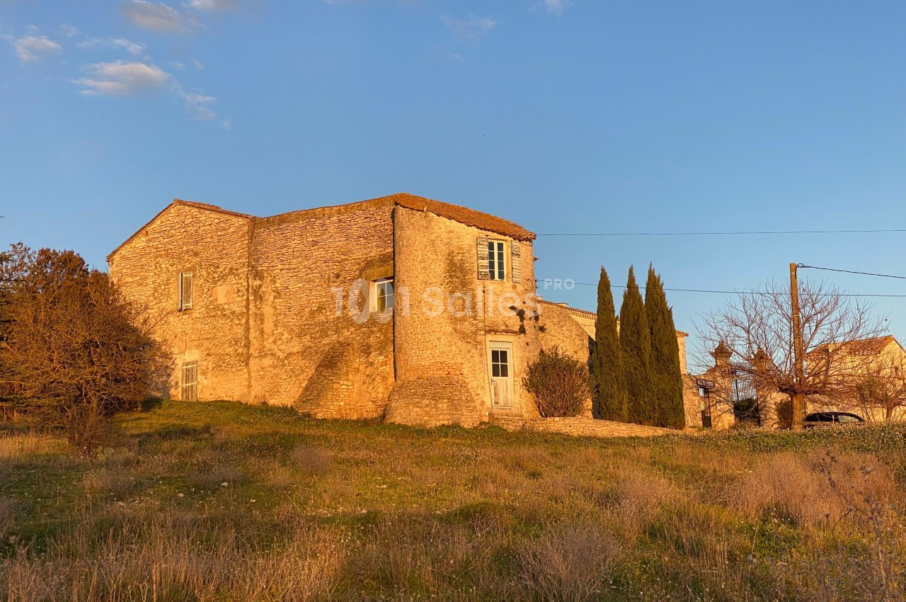 Maison en pierre éclairée par le soleil couchant, entourée de végétation et de quelques arbres.