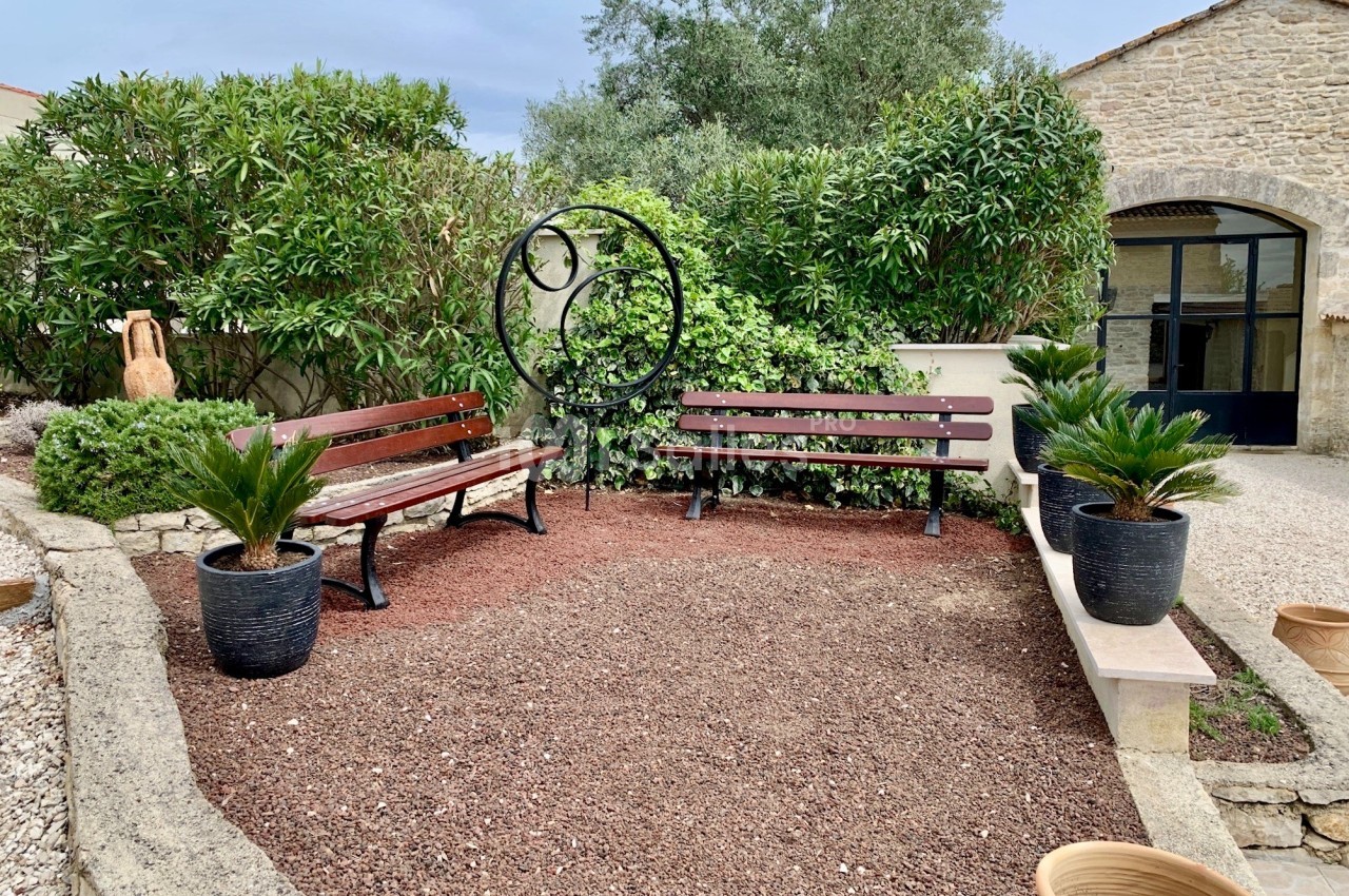 Deux bancs en bois entourés de plantes et de pots, disposés dans une cour avec un sol en gravier rouge.