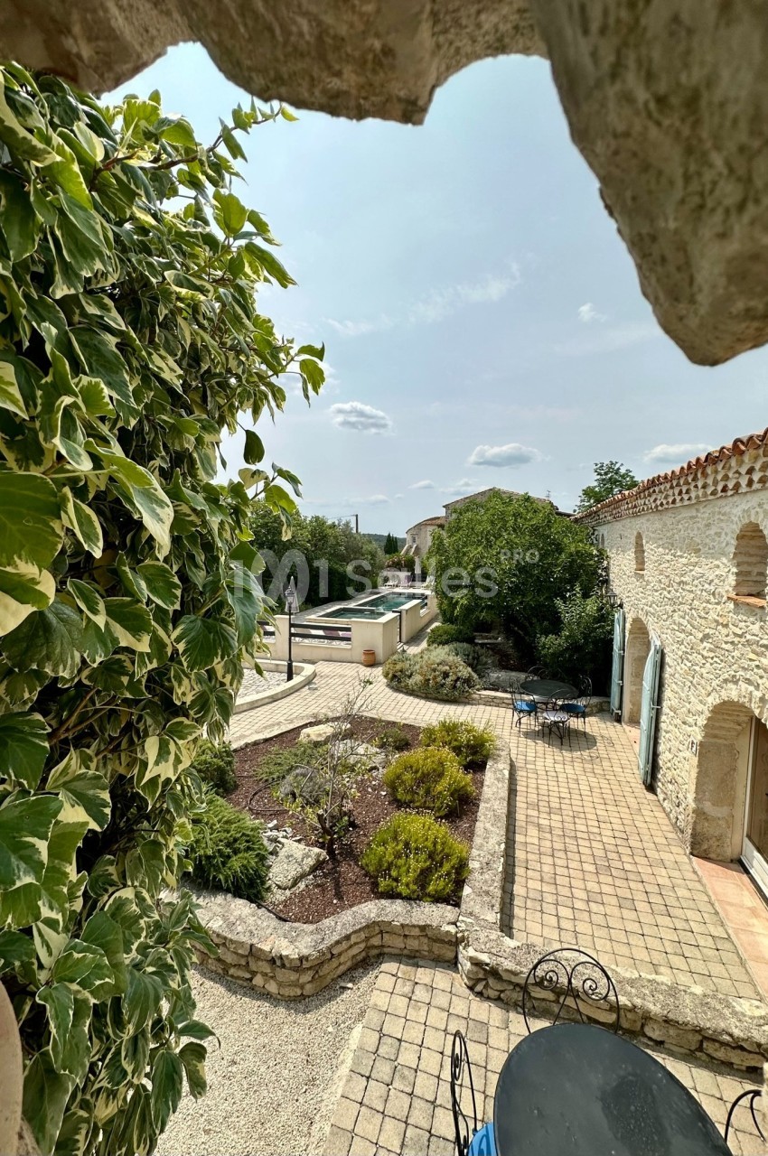 Vue d'une cour en pierre avec terrasse, végétation, piscine et ciel dégagé, encadrée par un mur en premier plan.