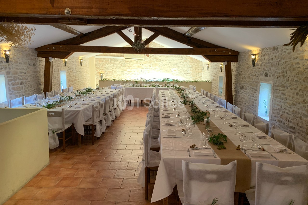 Salle de réception avec deux longues tables dressées, décorées de nappes blanches et de feuillages, dans un cadre en pierre.