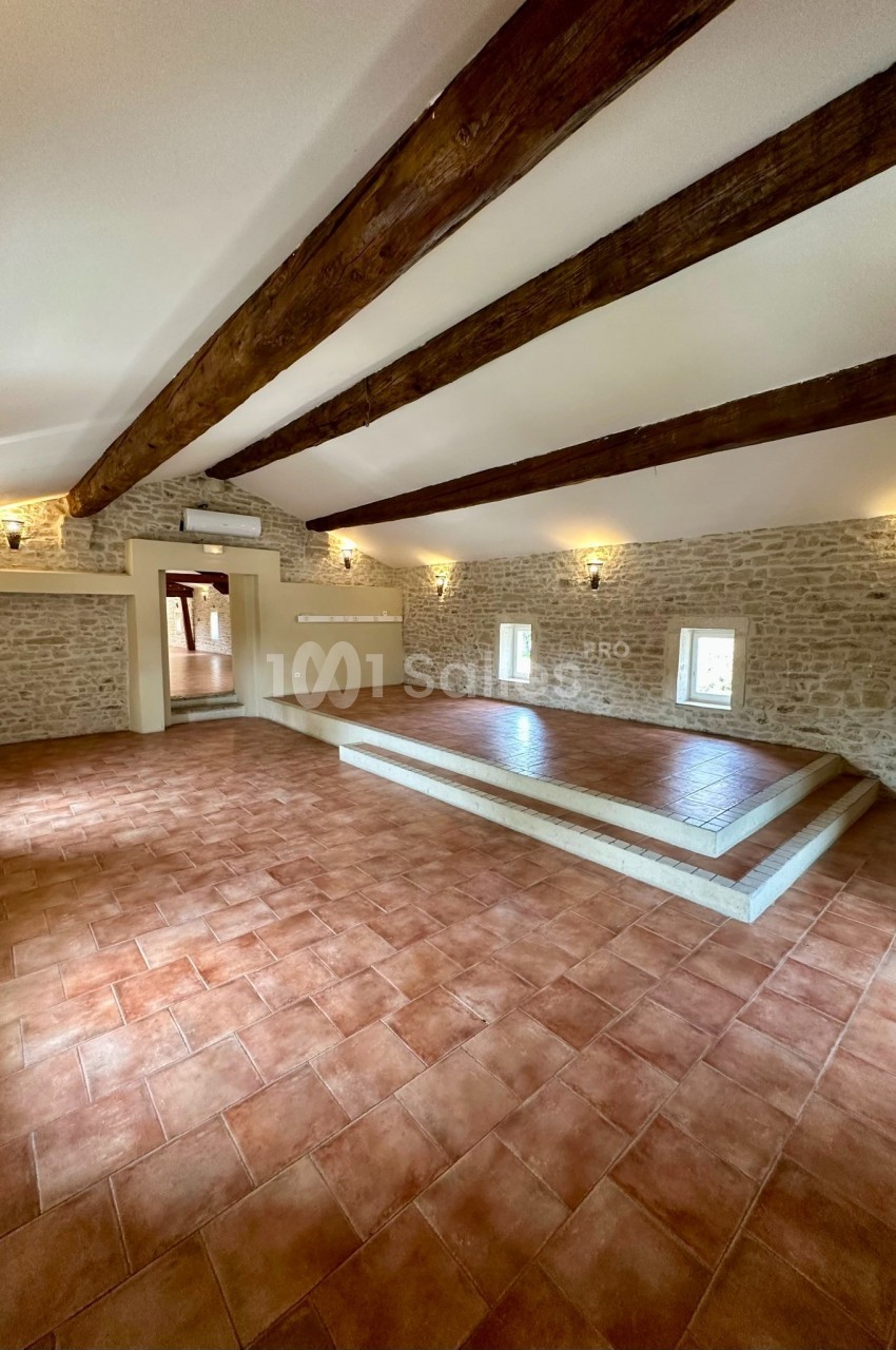 Salle lumineuse avec murs en pierre, poutres apparentes, sol en carrelage et estrade surélevée.
